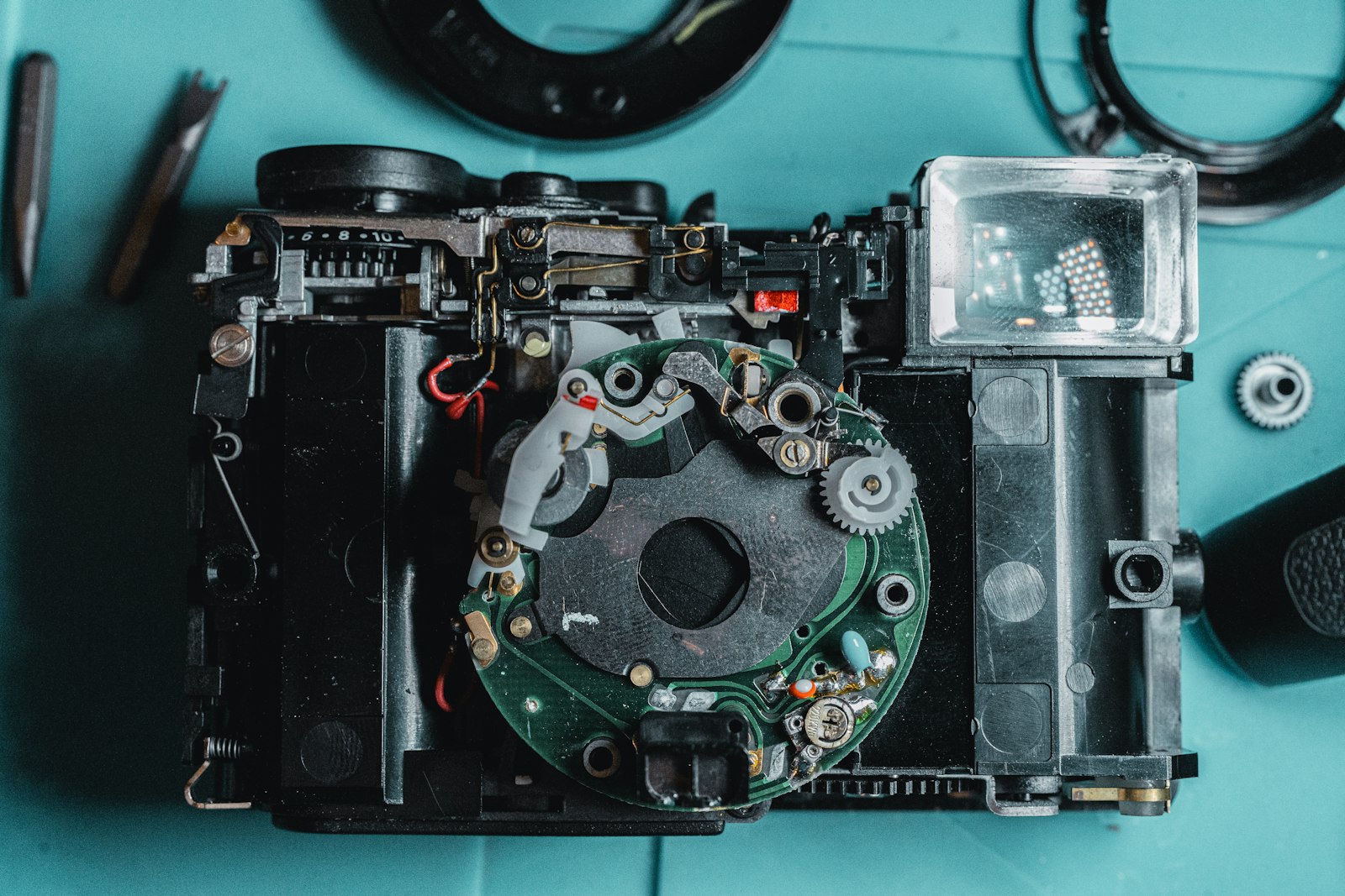 A camera stripped down to its internal circuit board and gears on a workbench, surrounded by loose components and tools