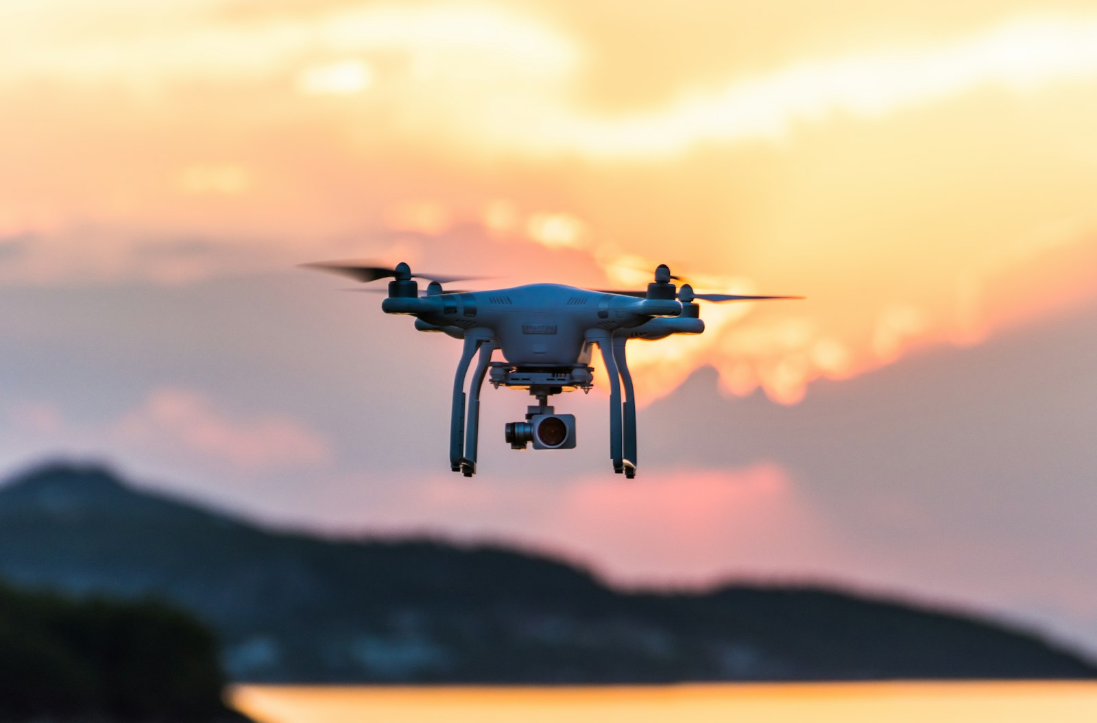 A white quadcopter drone with a gimbal-mounted camera hovering against an orange sunset sky, with hills and water blurred behind it