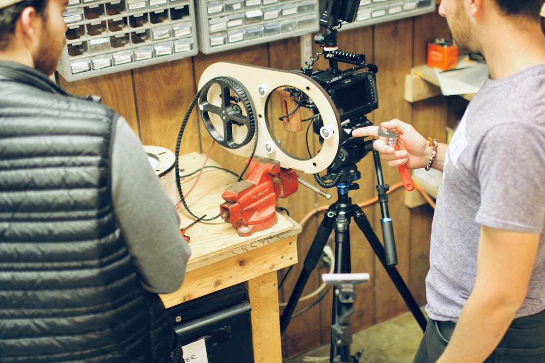 Two people examining a camera mounted on a tripod at a workshop bench, with parts bins on the wall behind