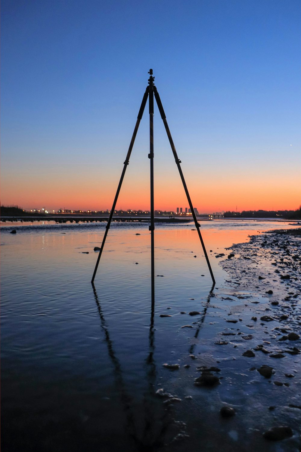An empty camera tripod standing in shallow water at sunset, with no camera mounted on it