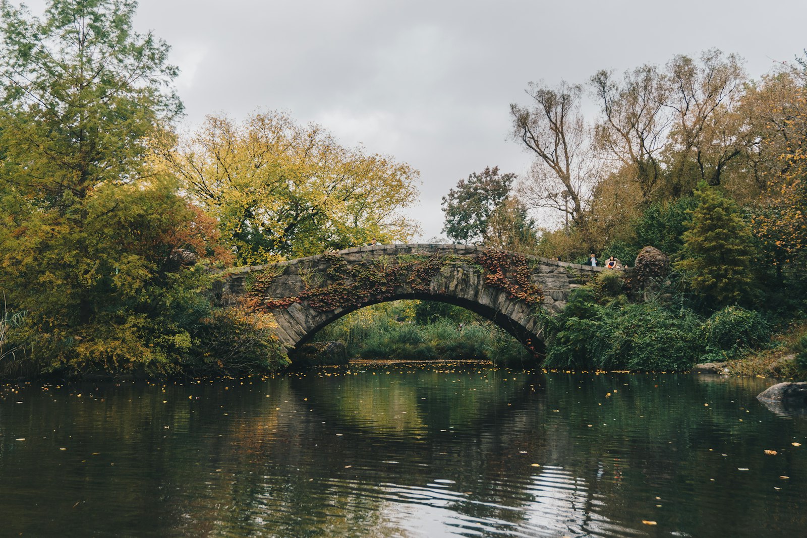 A stone arch bridge over a calm lake surrounded by autumn trees, with two people standing on the bridge
