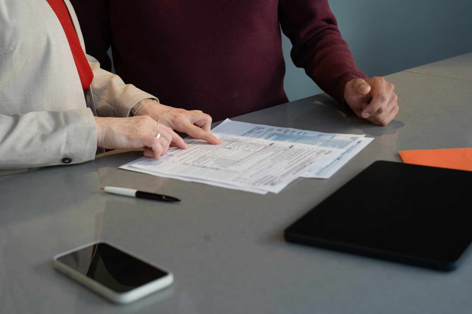Two people at a grey table going over a printed multi-page form, one pointing at a line on the page, with a pen, phone and tablet on the desk