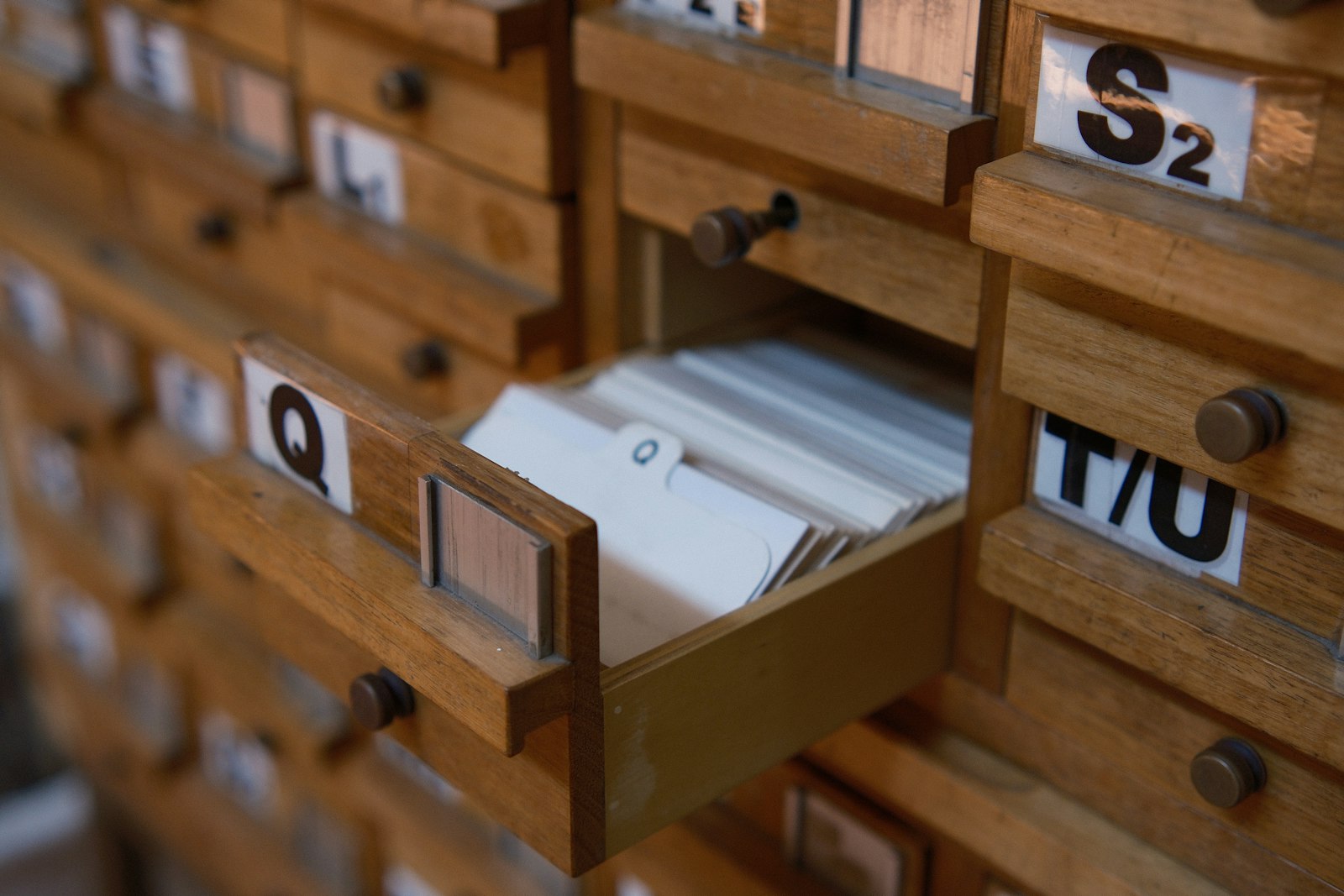 One open drawer of a wooden card catalogue packed with index cards, surrounded by closed drawers labelled with letters including Q, S and T slash U