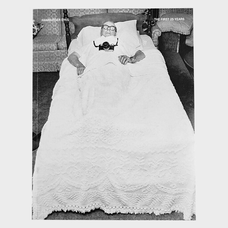 Cover of Hamburger Eyes – The First 25 Years, a black-and-white photo of a man in bed holding a camera to his chest