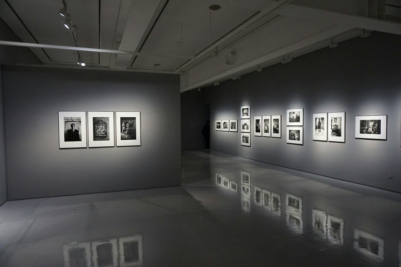Framed black and white photographs lit on the grey walls of an empty gallery, reflected in a polished concrete floor