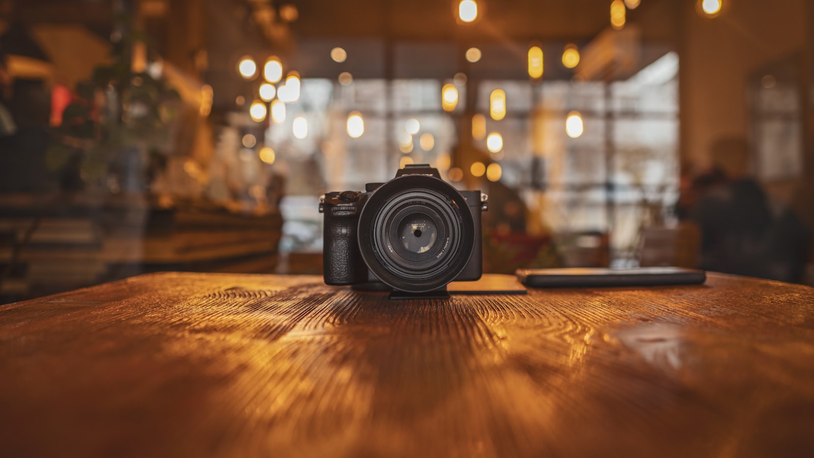 A single mirrorless camera body sitting alone on a wooden cafe table, warm bokeh lights behind it