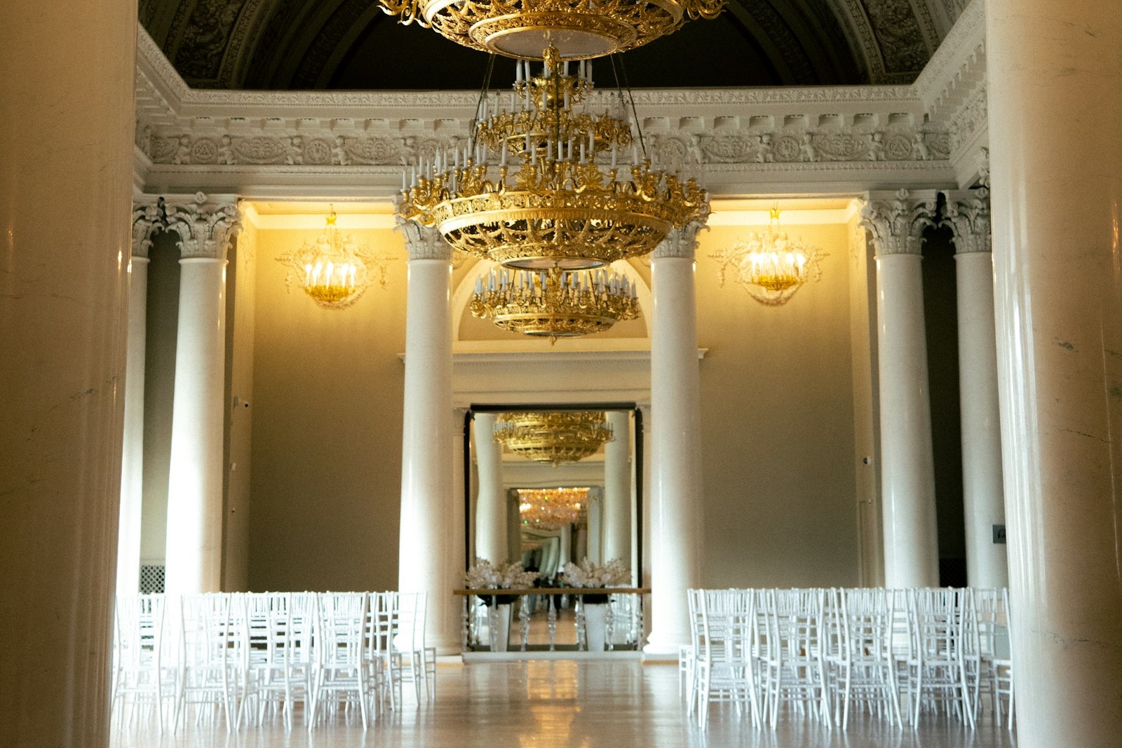 An empty ballroom with marble columns, gilded chandeliers and rows of white ceremony chairs set either side of a central aisle